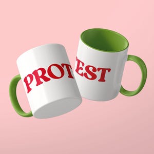 May include: Two white ceramic mugs with green handles and green interiors. The mug on the left features the red text PROT, while the mug on the right displays the red text EST. Both mugs have a smooth finish and a standard cylindrical shape.