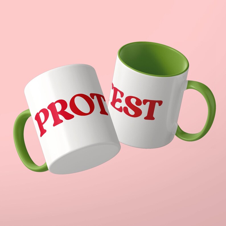 May include: Two white ceramic mugs with green handles and green interiors. The mug on the left features the red text PROT, while the mug on the right displays the red text EST. Both mugs have a smooth finish and a standard cylindrical shape.