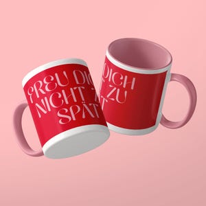 May include: Two ceramic mugs with a bright red exterior and a soft pink interior and handle. The mugs feature white text that reads "FREU DICH NICHT ZU SP&Auml;T" in a stylized, elegant font. The design is clean, with a smooth finish and a rounded handle.