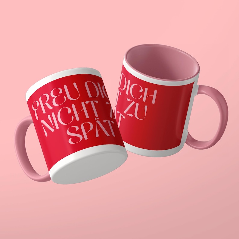 May include: Two ceramic mugs with a bright red exterior and a soft pink interior and handle. The mugs feature white text that reads "FREU DICH NICHT ZU SP&Auml;T" in a stylized, elegant font. The design is clean, with a smooth finish and a rounded handle.