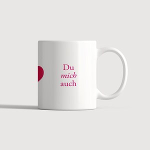 Pu&ograve; includere: Una tazza in ceramica bianca con un grafico a cuore rosso parziale sul lato sinistro e il testo magenta "Du mich auch" stampato in un carattere serif sulla parte anteriore. La tazza ha un corpo cilindrico standard e un manico curvo.