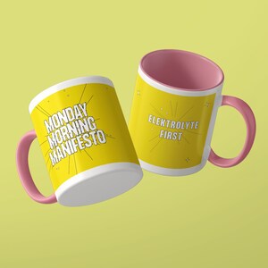 May include: Two ceramic mugs with pink handles and interiors. The yellow exterior of one mug features the black text MONDAY MORNING MANIFESTO with radiating lines. The other mug features the black text ELEKTROLYTE FIRST with radiating lines.
