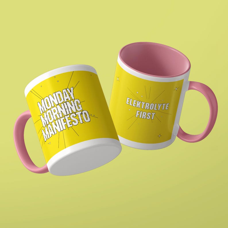 May include: Two ceramic mugs with pink handles and interiors. The yellow exterior of one mug features the black text MONDAY MORNING MANIFESTO with radiating lines. The other mug features the black text ELEKTROLYTE FIRST with radiating lines.