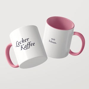 May include: Two white ceramic mugs with pink interiors and pink handles. One mug features the black text Lecker Kaffee, while the other mug displays the black text mit Schuss. The mugs are positioned at an angle against a plain background.