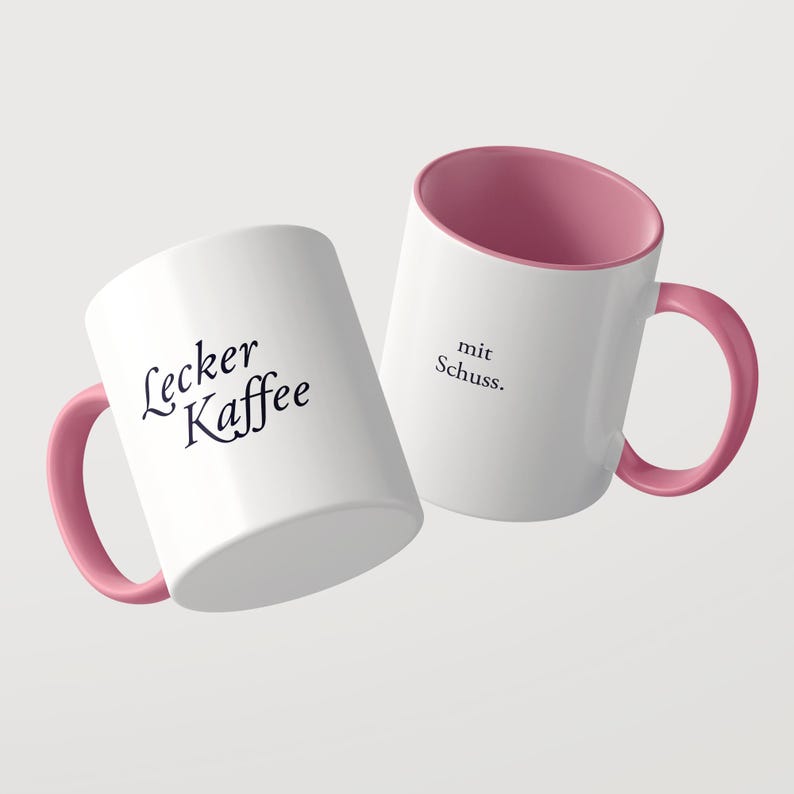 May include: Two white ceramic mugs with pink interiors and pink handles. One mug features the black text Lecker Kaffee, while the other mug displays the black text mit Schuss. The mugs are positioned at an angle against a plain background.