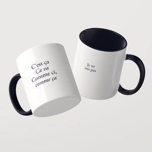 May include: Two white ceramic mugs with black interiors and black handles. The first mug features the black text: C'est &ccedil;a &Ccedil;a va Comme ci, comme &ccedil;a. The second mug features the black text: Je ne sais pas. Both mugs are displayed against a plain background.