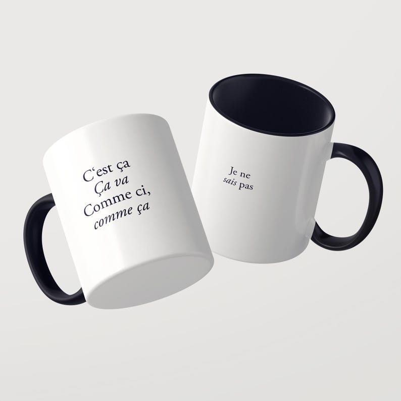 May include: Two white ceramic mugs with black interiors and black handles. The first mug features the black text: C'est &ccedil;a &Ccedil;a va Comme ci, comme &ccedil;a. The second mug features the black text: Je ne sais pas. Both mugs are displayed against a plain background.