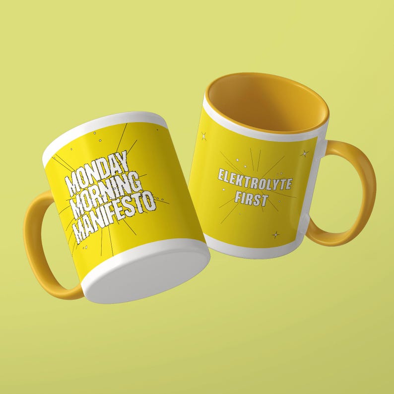 May include: Two yellow ceramic mugs with white interiors and yellow handles. One mug features the black text MONDAY MORNING MANIFESTO with radiating lines. The other mug displays the black text ELEKTROLYTE FIRST with small star accents.