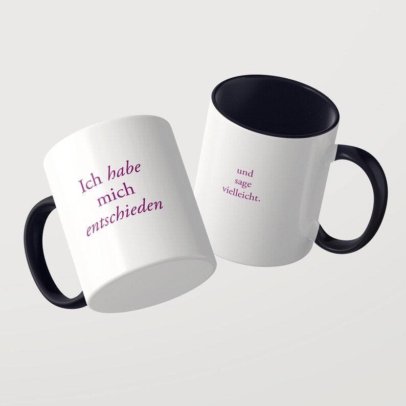 May include: Two white ceramic mugs with black handles and black interiors. The first mug features the purple text Ich habe mich entschieden, while the second mug displays the purple text und sage vielleicht. Both mugs have a smooth finish.