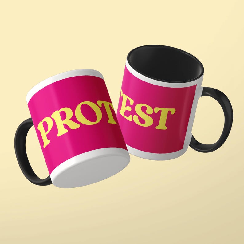 May include: Two ceramic mugs with black handles and interiors. The exterior of each mug is bright pink with a white border. The yellow text PROTEST is printed across the mugs, with PROT on one mug and EST on the other, in a bold serif font.