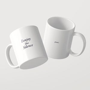 May include: Two white ceramic mugs with black text. One mug features the phrase "I enjoy the Silence" in a serif font. The second mug displays the word "pssst." in a smaller, simple font. Both mugs have a standard handle and cylindrical shape.