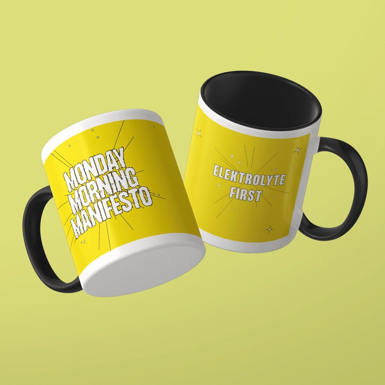 May include: Two ceramic mugs with black handles and interiors. The exterior is yellow with white text. One mug features the text MONDAY MORNING MANIFESTO with radiating lines. The other mug displays the text ELEKTROLYTE FIRST with small star accents.