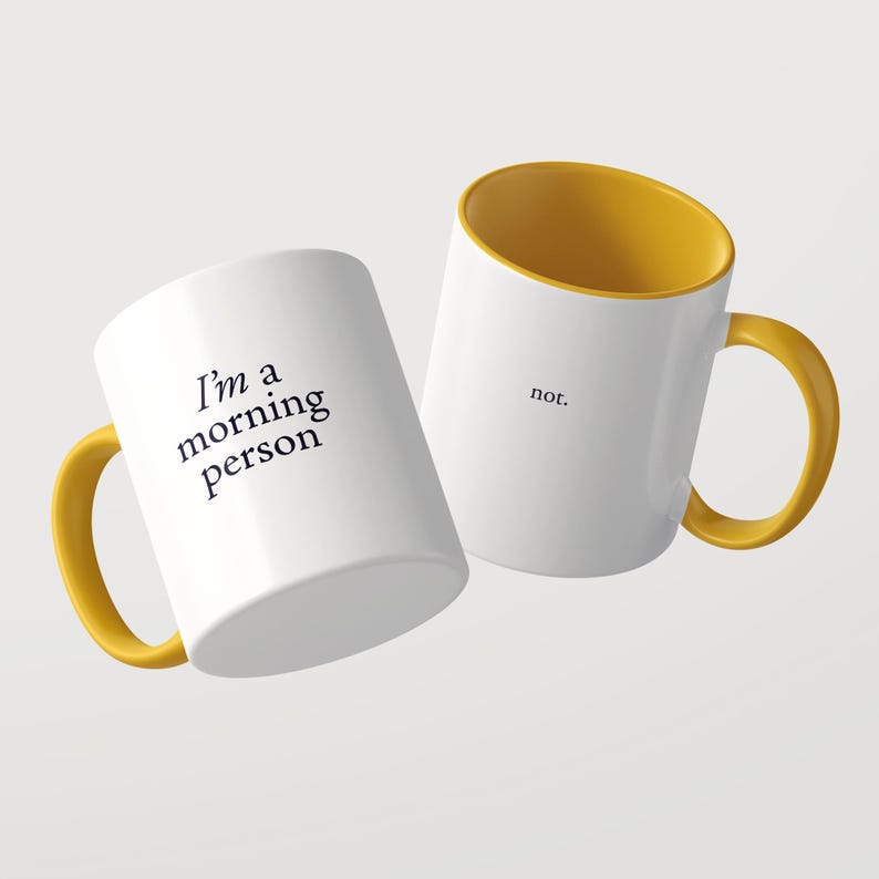 Może przedstawiać: Dwa białe ceramiczne kubki z musztardowo-ż&oacute;łtym wnętrzem i uchwytem. Jeden kubek zawiera czarny napis 'I'm a morning person', a drugi czarny napis 'not.' prostą czcionką szeryfową, przedstawione na jednolitym tle.