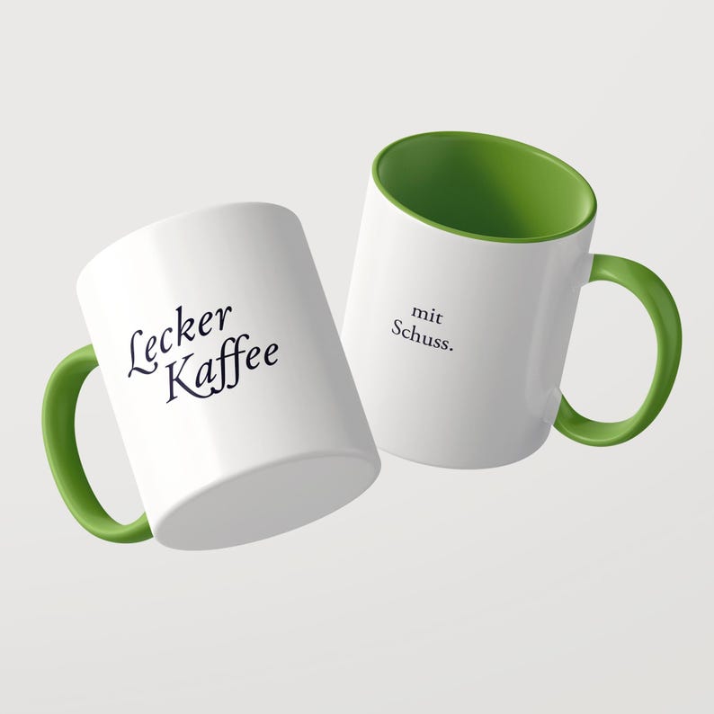 May include: Two white ceramic mugs with green interiors and green handles. One mug features the black text Lecker Kaffee, while the other mug displays the black text mit Schuss. The mugs are positioned at an angle against a plain background.