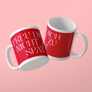 May include: Two white ceramic mugs with a bright red exterior finish. The mugs feature white text printed in a stylized font that reads "FREU DICH NICHT ZU SP&Auml;T". Each mug has a curved handle on the side and a plain white interior surface.
