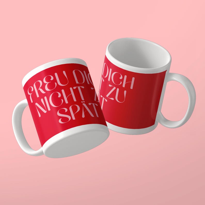 May include: Two white ceramic mugs with a bright red exterior finish. The mugs feature white text printed in a stylized font that reads "FREU DICH NICHT ZU SP&Auml;T". Each mug has a curved handle on the side and a plain white interior surface.