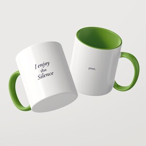 May include: Two white ceramic mugs with green interiors and green handles. One mug features the black text 'I enjoy the Silence' and the other mug features the black text 'pssst.' printed on the exterior surface of the white ceramic body.