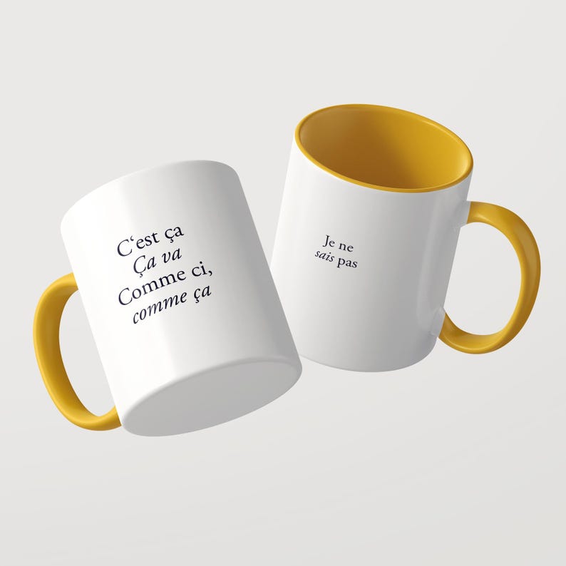 May include: Two white ceramic mugs with mustard yellow interiors and handles. The left mug features the black text C'est &ccedil;a &Ccedil;a va Comme ci, comme &ccedil;a. The right mug displays the black text Je ne sais pas on its side. Both mugs are cylindrical.