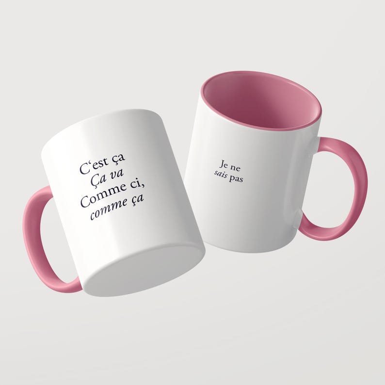 May include: Two white ceramic mugs with pink interiors and matching pink handles. The first mug features the black text C'est &ccedil;a &Ccedil;a va Comme ci, comme &ccedil;a. The second mug displays the black text Je ne sais pas on its exterior surface.