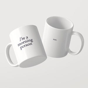 Może przedstawiać: Dwa białe kubki ceramiczne z czarnym tekstem. Jeden kubek zawiera napis 'I'm a morning person' czcionką szeryfową. Drugi kubek, umieszczony nieco za nim i po prawej stronie, zawiera słowo 'not.' wykonane tą samą czarną czcionką szeryfową.