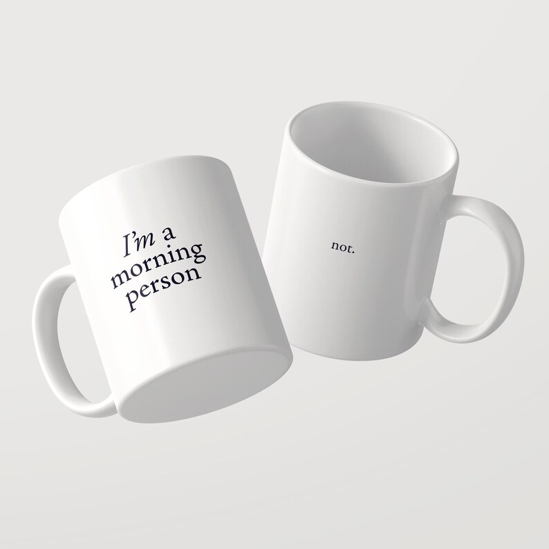 Może przedstawiać: Dwa białe kubki ceramiczne z czarnym tekstem. Jeden kubek zawiera napis 'I'm a morning person' czcionką szeryfową. Drugi kubek, umieszczony nieco za nim i po prawej stronie, zawiera słowo 'not.' wykonane tą samą czarną czcionką szeryfową.
