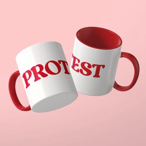 May include: Two white ceramic mugs with red interiors and red handles. The mug on the left features the red text PROT, while the mug on the right features the red text EST. Both mugs have a smooth, glossy finish and a standard cylindrical shape.