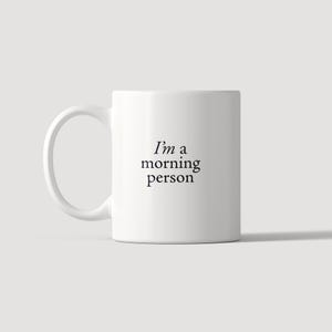 Może przedstawiać: Biały ceramiczny kubek z uchwytem po lewej stronie, z czarnym nadrukiem "I'm a morning person" wyśrodkowanym na przedniej części. Kubek ma gładki, cylindryczny korpus i prosty design, przeznaczony do serwowania gorących napoj&oacute;w.