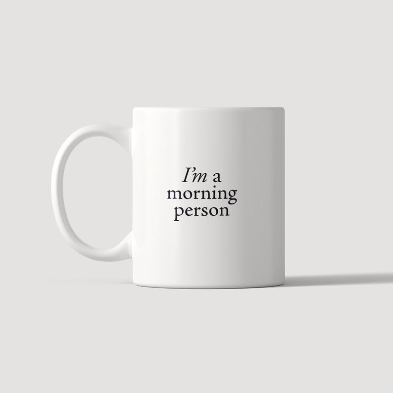 Może przedstawiać: Biały ceramiczny kubek z uchwytem po lewej stronie, z czarnym nadrukiem "I'm a morning person" wyśrodkowanym na przedniej części. Kubek ma gładki, cylindryczny korpus i prosty design, przeznaczony do serwowania gorących napoj&oacute;w.