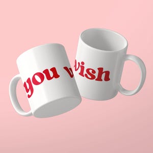 Può includere: Due tazze in ceramica bianca con il testo rosso you wish stampato in un carattere grassetto e arrotondato. Le tazze sono posizionate ad angolo, mostrando il manico e il corpo cilindrico di ogni recipiente su uno sfondo rosa chiaro.
