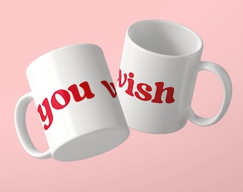 May include: Two white ceramic mugs featuring the red text you wish printed in a bold, rounded font. The mugs are positioned at an angle, showcasing the handle and the cylindrical body of each vessel against a solid light pink background.