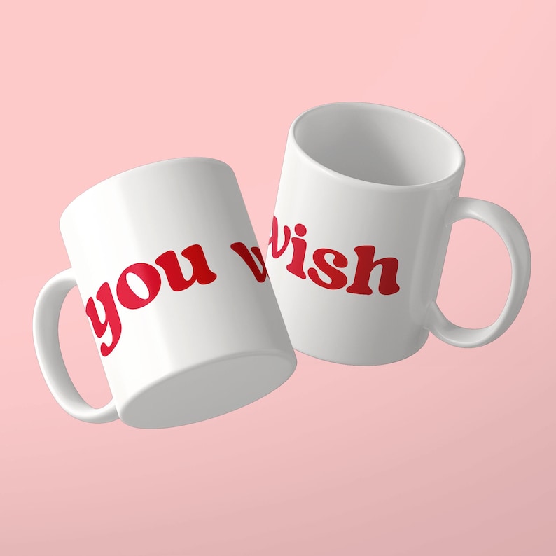 May include: Two white ceramic mugs featuring the red text you wish printed in a bold, rounded font. The mugs are positioned at an angle, showcasing the handle and the cylindrical body of each vessel against a solid light pink background.