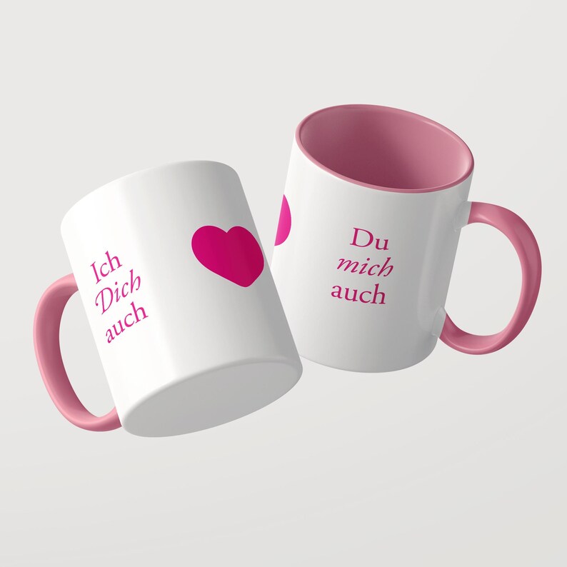 Pu&ograve; includere: Due tazze in ceramica bianca con interno e manico rosa. Una tazza presenta un cuore rosa e il testo Ich Dich auch in rosa. La seconda tazza mostra il testo Du mich auch in rosa. Le tazze sono posizionate ad angolo su uno sfondo a tinta unita.