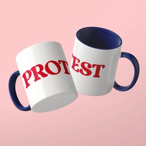 May include: Two white ceramic mugs with dark blue interiors and handles. The mug on the left features the red text PROT, while the mug on the right displays the red text EST. Both mugs have a smooth, glossy finish and a standard cylindrical shape.