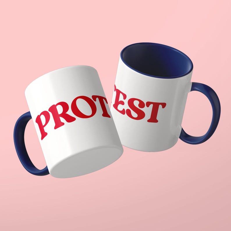 May include: Two white ceramic mugs with dark blue interiors and handles. The mug on the left features the red text PROT, while the mug on the right displays the red text EST. Both mugs have a smooth, glossy finish and a standard cylindrical shape.