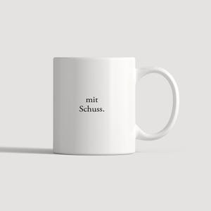 May include: A white ceramic mug featuring a side handle and the black printed text "mit Schuss." centered on the front. The mug has a smooth, cylindrical body with a clean, minimalist design and a standard circular rim at the top.