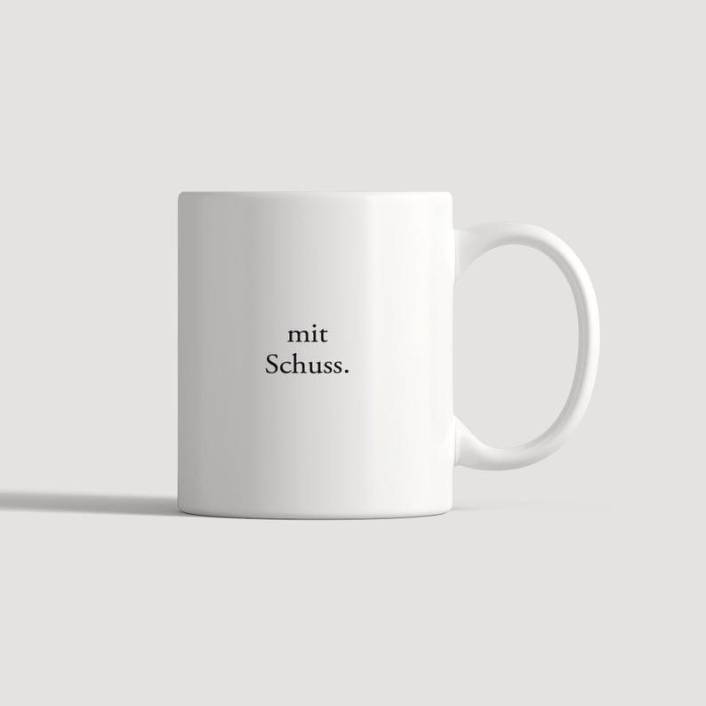 May include: A white ceramic mug featuring a side handle and the black printed text "mit Schuss." centered on the front. The mug has a smooth, cylindrical body with a clean, minimalist design and a standard circular rim at the top.