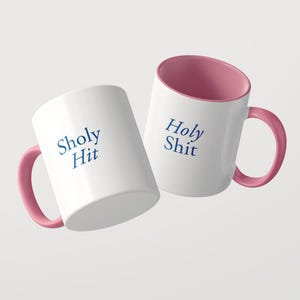 May include: Two white ceramic mugs with pink interiors and pink handles. One mug features the blue text Sholy Hit, while the other mug displays the blue text Holy Shit. The mugs are positioned at an angle against a plain light background.
