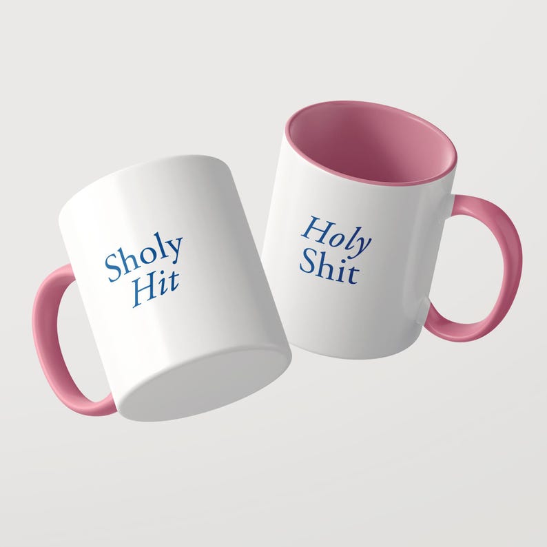 May include: Two white ceramic mugs with pink interiors and pink handles. One mug features the blue text Sholy Hit, while the other mug displays the blue text Holy Shit. The mugs are positioned at an angle against a plain light background.