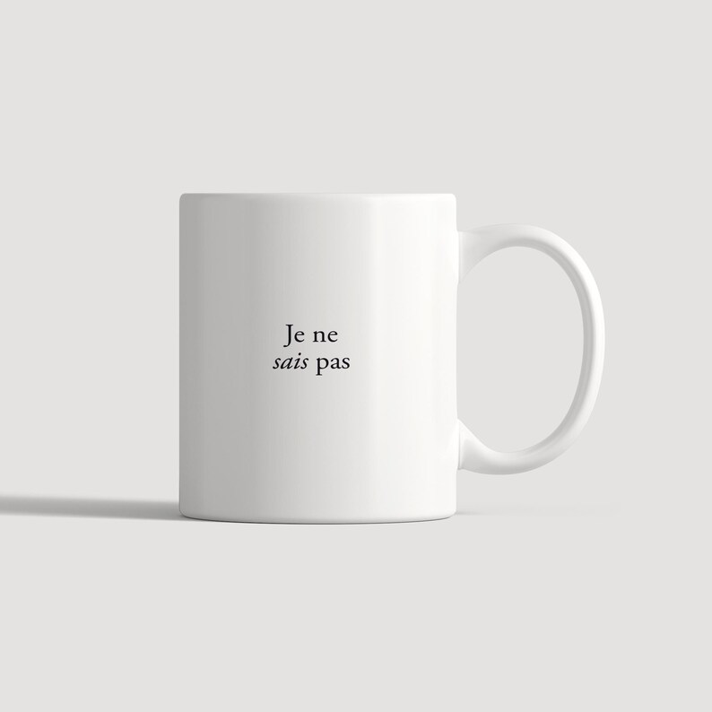 May include: A white ceramic mug featuring a side handle and the black printed text Je ne sais pas centered on the front. The mug has a smooth, cylindrical body and a clean, minimalist design suitable for holding hot or cold beverages.