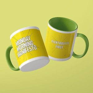 May include: Two ceramic mugs with green handles and interiors. The left mug features yellow sides with the black text MONDAY MORNING MANIFESTO. The right mug has yellow sides with the black text ELEKTROLYTE FIRST. Both mugs feature thin black line graphics.
