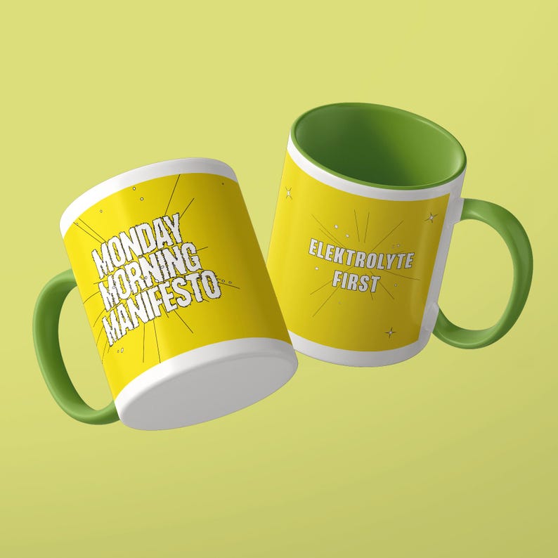 May include: Two ceramic mugs with green handles and interiors. The left mug features yellow sides with the black text MONDAY MORNING MANIFESTO. The right mug has yellow sides with the black text ELEKTROLYTE FIRST. Both mugs feature thin black line graphics.