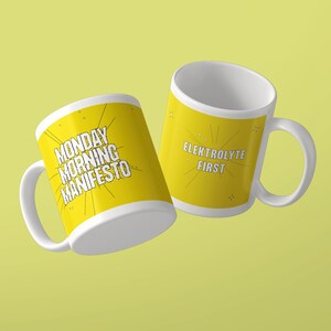 May include: Two white ceramic mugs with yellow exterior surfaces. One mug features the black text MONDAY MORNING MANIFESTO with radiating lines. The other mug displays the white text ELEKTROLYTE FIRST with radiating lines and small star accents.