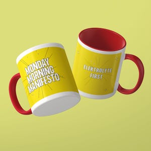 May include: Two ceramic mugs with bright yellow exteriors and red interiors and handles. One mug features the black text MONDAY MORNING MANIFESTO with radiating lines. The other mug displays the black text ELEKTROLYTE FIRST with star accents.