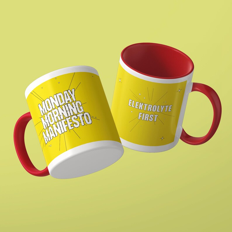 May include: Two ceramic mugs with bright yellow exteriors and red interiors and handles. One mug features the black text MONDAY MORNING MANIFESTO with radiating lines. The other mug displays the black text ELEKTROLYTE FIRST with star accents.