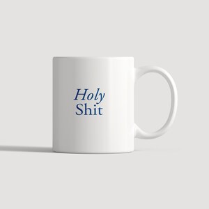 May include: A white ceramic mug featuring the text Holy Shit printed in a dark blue serif font on the front. The mug has a smooth cylindrical body and a curved handle on the right side, providing a simple and functional design for daily beverage use.