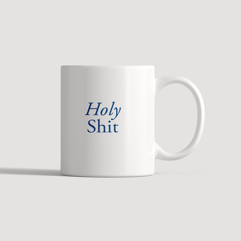 May include: A white ceramic mug featuring the text Holy Shit printed in a dark blue serif font on the front. The mug has a smooth cylindrical body and a curved handle on the right side, providing a simple and functional design for daily beverage use.