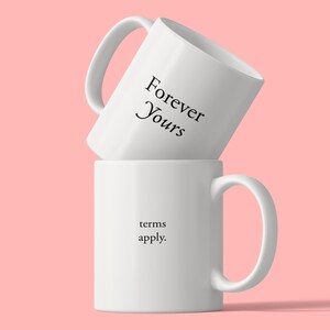 May include: Two white ceramic mugs stacked vertically. The top mug is tilted and features the black text Forever Yours. The bottom mug is upright and features the black text terms apply. Both mugs have a smooth finish and a single side handle.
