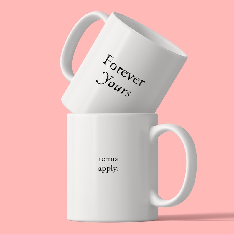 May include: Two white ceramic mugs stacked vertically. The top mug is tilted and features the black text Forever Yours. The bottom mug is upright and features the black text terms apply. Both mugs have a smooth finish and a single side handle.