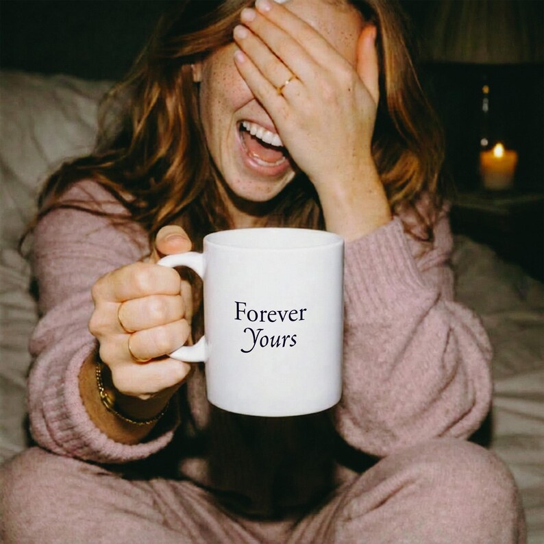 May include: A white ceramic mug with a handle, featuring the black printed text Forever Yours centered on the front. The mug is held by a person wearing a mauve long-sleeved sweater, with the person's face partially obscured by their hand.