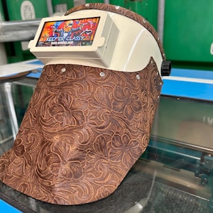 May include: A welding helmet with a cream-coloured shell and a brown, floral-patterned leather face shield. The helmet features a rectangular screen with the text "KEEP ER CLASSY" and a colourful graphic. The face shield is adorned with rivets.